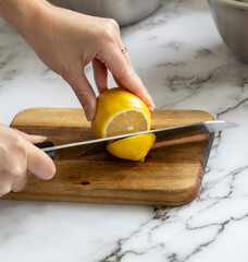 Petite planche à découper en bois sur une surface de marbre avec main qui tient un couteau en métal à lame droite et manche noir en train de couper le bout d'un citron jaune mur dans la cuisine