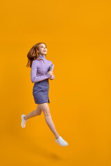 Fototapeta na wymiar Optimistic redhead lady at weekend jumping, hold notebooks in hands on way home, in air, wearing casual shirt and skirt isolated yellow orange background. Side view, copy space. Full-length portrait