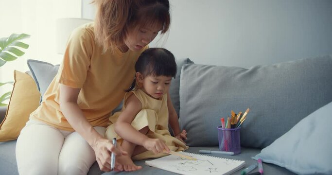 Happy cheerful Asia family mom teach girl paint use album and colourful pencils having fun relax on couch in living room at house. Spending time together, Social distance, Quarantine for coronavirus.