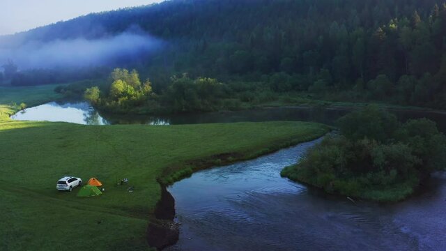 Camping by the river. Aerial sunrise view of the wild campsite by the river. Drone view of the campers with car and tents set on the green meadow close to the river