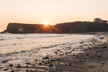 A bright orange sun sets over the ocean and a rocky seashore