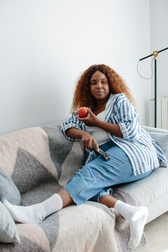 Woman reclined on the couch eating apple holding tv remote
