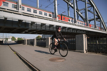 Morning training of female cyclist riding along the embankment under the railway bridge with a train at sunset. Sports in the city. Sports equipment.
