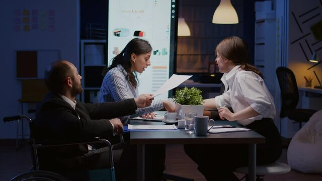 Overworked paralyzed disabled entrepreneur man in wheelchair sitting at desk overworking in company office meeting room. Manager is ignored while coworkers analyzing statistics paperwork late at night