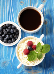 A cup of coffee and Cottage cheese in white bowl with raspberries and blueberries on wooden background.