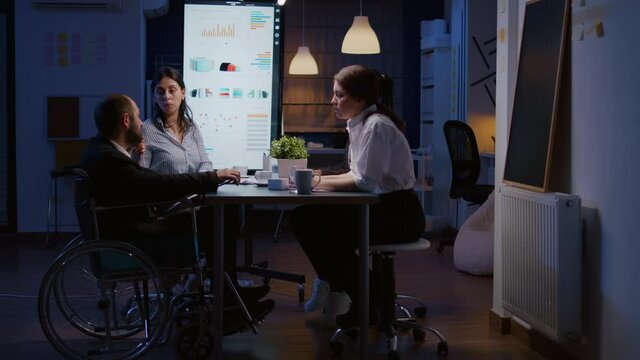 Overworked disabled businessman in wheelchair is ignored while working in company business office meeting room late at night. Workaholics multi-ethnic businesspeople brainstorming strategy ideas