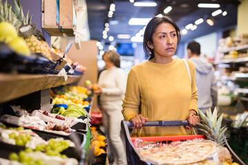 Pensive latina buying groceries at store, walking with shopping cart among shelves and choosing products