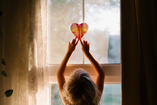 A little girl putting a paper heart against the window at the sunrise