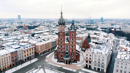Aerial view of the Krakow’s Rynek Głowny (Central Square) surrounded by historic buildings. Twin towers of the Basilica of Saint Mary against clear white sky in the background. City Skyline.