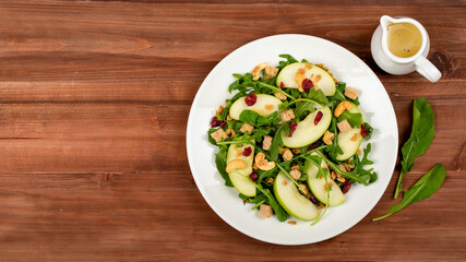 Green apple slice salad with rocket, oat granular, dried cranberry, and cashew nut placed in white dish on wooded table. Oil vinegar salad dressing, a side dish. Two rocket leaf used as decoration