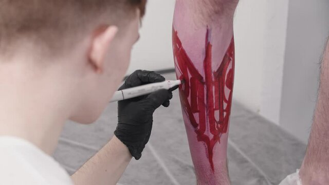 Young man tattoo artist drawing a sketch with letters on the leg on his client with a red marker