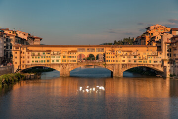 Florence, Italy -20 June, 2019 : view of Ponte Vecchio Bridge glittering in sunset sun rays , river Arno and residential area of the city at golden hour.