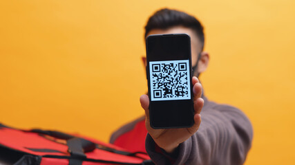Delivery man holding a red parcel box. Showing mobile phone screen in the camera with QR code. Concept of parcel delivery and digital pay or E-Wallet. Yellow background studio shot.