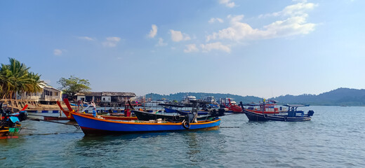 Obraz premium Scenic tropical fishing village with colorful traditional wooden boats anchored in calm blue sea, peaceful coastal lifestyle with clear sky and lush island background, idyllic maritime scenery