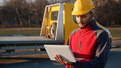 Young engineer wearing a uniform and a safety helmet standing with a yellow truck behind him....