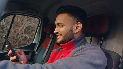 Young delivery guy sitting in the front seat of the van with hand on steering. Holding a phone in his other hand and smiling. Using his phone. Wearing a red uniform. Courier concept.  © CameraCraft