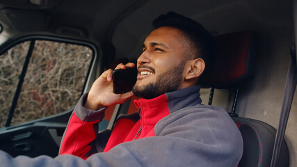 Delivery boy driving a van and talking on phone. Holding a phone in his hand. Dressed in red uniform. Smiling and talking to someone on call. Young India Guy. Parcel delivery concept.