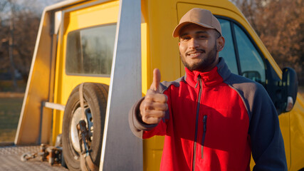 Worker wearing cap and uniform with a yellow truck behind him. Boy smiling and making a thumbs up sign. Concept of automobile mechanic. Spare tire can be seen in the truck behind.