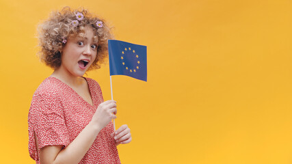Beautiful young girl holding a small European Union Flag in her hand. Astonished girl wearing casual pink top. Studio shot isolated on yellow background. Symbol of Europe, EU association. 
