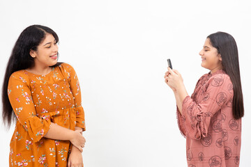 Two young Indian girls taking photo on their smart phone. Cute teenagers having fun with each other on a white background.