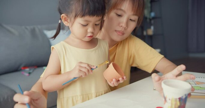 Happy cheerful Asia family mom teach toddler girl paint ceramic pot having fun relax on table in living room at house. Spending time together, Social distance, Quarantine for coronavirus prevention.