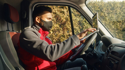 Indian guy sitting on the driving seat of the van using a laptop. Dressed in red zipper wearing a black Mask. Concept of car repair and maintenance. Sunlight hitting his face.  © CameraCraft