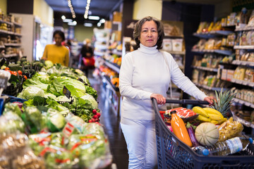 Mature positive woman choosing food products on shelves in grocery shop