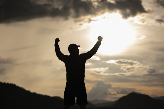 Silhouette Young man raised hand in the air with success goal on sunset background. Man standing on top of the mountain freedom life concept.