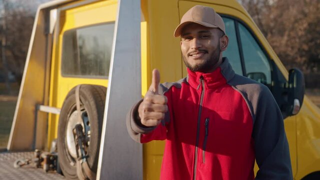 Young Indian mechanic wearing a cap smiling and making thumbs up sign at the camera. Yellow truck can be seen behind him with a spare tire. Concept of automobile mechanics. Closeup footage. 