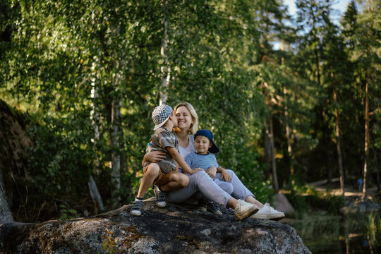 young beautiful woman enjoying spending time with two kids. Elder son kissing mom on cheek. Family is sitting on big rock in Mon Repos Park, Vyborg. Image with selective focus