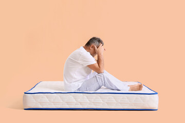 Sad mature man sitting on soft mattress against color background