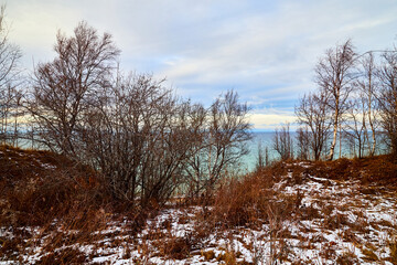 Nature landscape with dramatic sky and clouds, lake with ice, shore with snow and dark trees without leaves in cold winter or autumn day or evening
