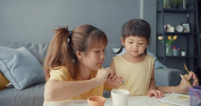 Happy cheerful Asia family mom teach toddler girl paint ceramic pot having fun relax on table in living room at house. Spending time together, Social distance, Quarantine for coronavirus prevention.