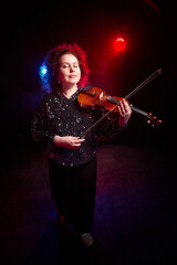 A brunette curly girl in black shiny clothes and with violin on a black background. Model, actress...