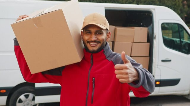 Delivery boy carrying a cardboard box on his shoulder. Smiling and making thumbs up sign at the camera. A white van with stock of cardboard boxes in the background. Concept of parcel delivery service.