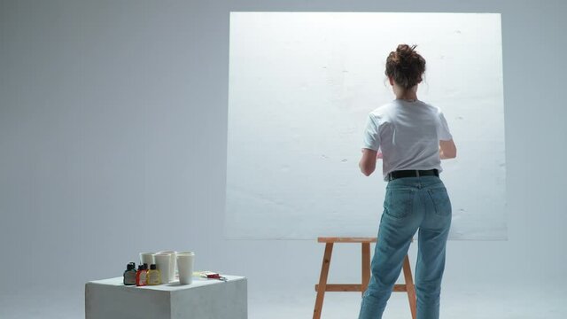 Back view, female artist applies a layer of white primer on a large canvas in a white room, a talented artist preparing to draw.
