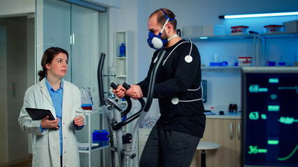 Performance athlete talking with medical researcher running on on cross trainer in sports science laboratory measuring VO2 max, heart rate, psychological resistance and muscular endurance. Monitor