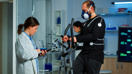 Science sport doctor asking patient about his health while sportsman running on cross trainer with mask and electrodes attached on body monitoring vo2. Woman researcher measuring endurance of patient.