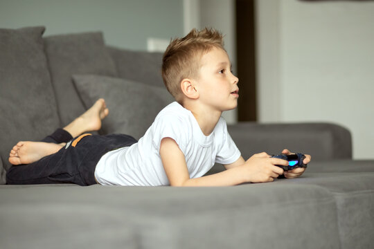 A child with a joystick in his hands lies on the couch at home and is intently playing the console