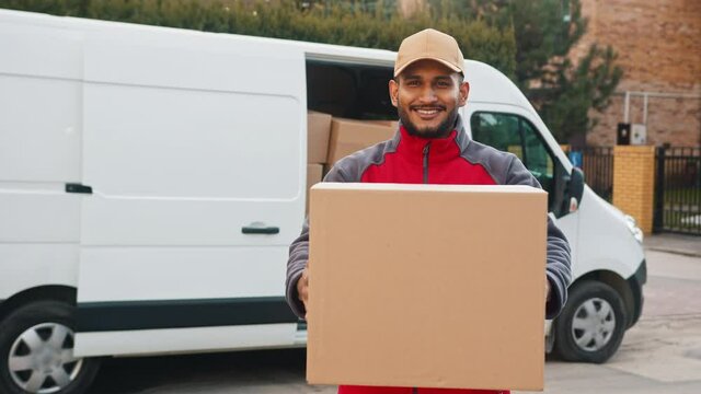 Delivery boy holding a cardboard box in his hands. Smiling and moving the box towards the camera. A white van with stock of cardboard boxes in the background. Concept of parcel delivery service. 