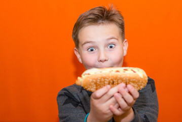 Surprised 10s boy wants to bite a big hot dog.Closeup orange studio background.