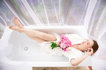 Beautiful girl in a white bathrobe with a bouquet of pink peonies near the bath. © nagaets