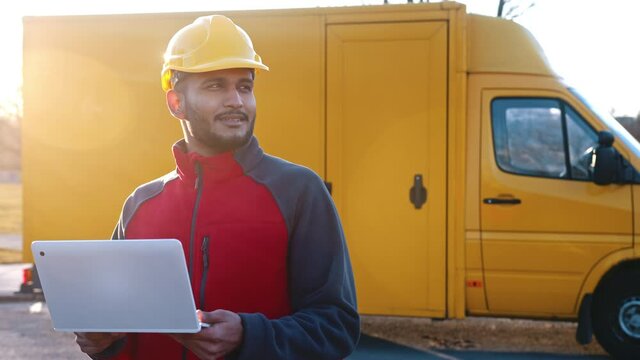A Young Indian engineer wearing safety yellow helmet holding a laptop in his hands. Checking his planned work looks satisfied. A yellow van can be seen in the background. 