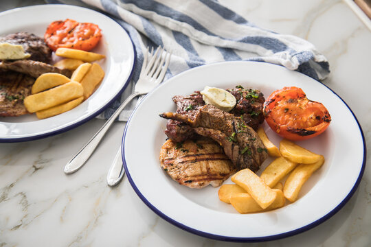 Grillteller mit Rindersteak, H&uuml;hnchen Brustfilet, Lamm Filet dazu Steakhaus Pommes, Grilltomate und Kr&auml;uterbutter auf Emaille Teller blau wei&szlig;, Tuch und Marmor Hintergrund hell 