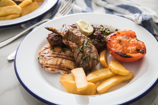 Grillteller mit Rindersteak, H&uuml;hnchen Brustfilet, Lamm Filet dazu Steakhaus Pommes, Grilltomate und Kr&auml;uterbutter auf Emaille Teller blau wei&szlig;, Tuch und Marmor Hintergrund hell 