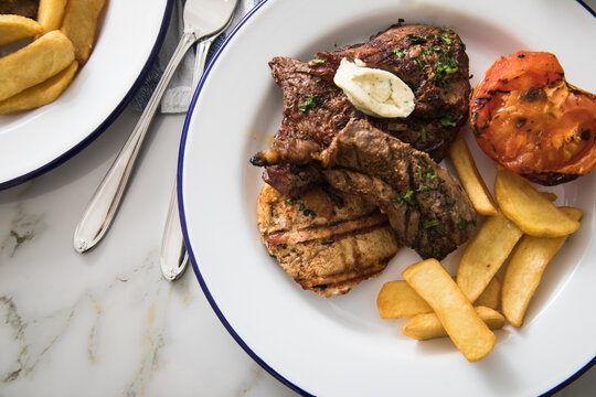 Grillteller mit Rindersteak, H&uuml;hnchen Brustfilet, Lamm Filet dazu Steakhaus Pommes, Grilltomate und Kr&auml;uterbutter auf Emaille Teller blau wei&szlig; und Marmor Hintergrund hell 