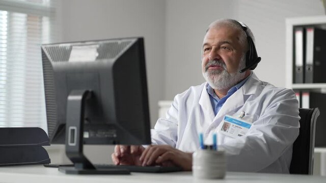 Grey male doctor remotely video communications with customer and records medical history in computer