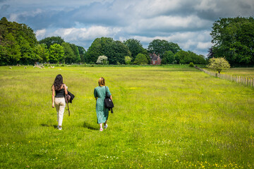 A blonde girl in a green dress and a brunette in white pants walk through a prairie full of yellow flowers towards the farmhouse visible at the back