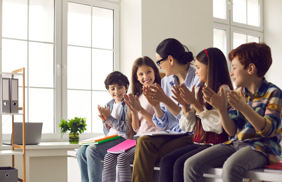Young female teacher together with group of happy children clapping hands while sitting in classroom. Educator and junior high students have fun in school. Concept of education, school and learning.