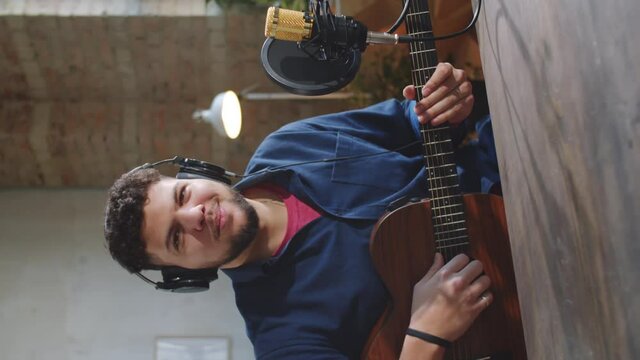 Vertical shot of young male musician in headphones waving at camera, talking into mic and showing how to play the guitar while giving online class in recording studio